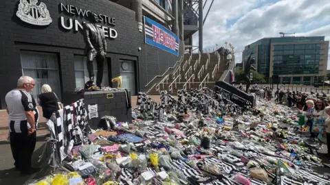 Andy Greenwood/BBC Hundreds of Newcastle United shirts, flowers, cards and other tributes on the floor outside St James' Park in Newcastle. Dozens of people are lined up looking at the tributes to Kevin Keegan. There is a blue and white banner hanging from the side of the stadium, between the Newcastle United sign and the steps, with the words The Messiah, in tribute to Kevin Keegan.