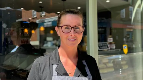 A woman looks straight at the camera. She is wearing glasses and a dark grey top. You can see the start of an apron. In the background is the window of the shop.