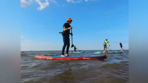 SUP Lincs A man wearing a red helmet on a red paddleboard on the sea, with other people on paddleboards in the background.