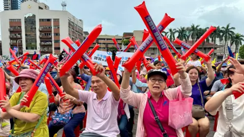 BBC/Tessa Wong Crowd The crowd sat at New Taipei's daytime anti-recall rally lifted large red inflatable rods and crossed them to show that they opposed the big recall. In the foreground, you can see a woman wearing a pink polo shirt and a pink cardigan with a pink bag and a man wearing a lightweight polo shirt. In the background, you can see municipal buildings and trees.