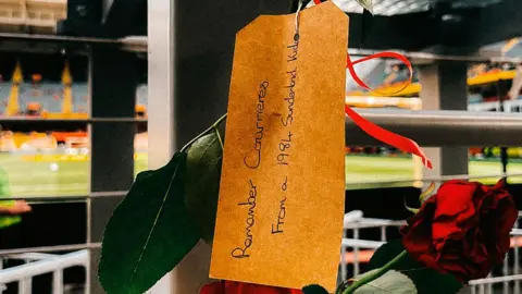 RC Lens A red rose is attached to a stadium railing with a brown memorial tag hanging from its stem. The handwritten tag reads, “Remember Courage. From a 1984 Sunderland Kid.” Blurred stadium seating and a football pitch are visible in the background.