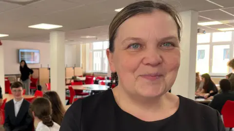 Teacher from Brook Mead Academy, in Leicester, looking at the camera with some students in the background.