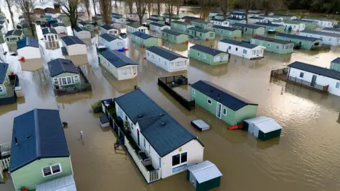 PA Media Green and white static caravans, which are dotted between trees, are seen deluged by brown floodwater which is at door height