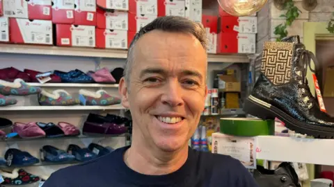 Declan Hughes is smiling. He is wearing a black jumper and standing beside a large black shoe on a white platform. A number of shoes and shoe boxes are visible behind him. 