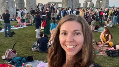 Alicia Moseley A selfie of Alicia Mosley at Stonehenge. She has long brown hair and is smiling while looking at the camera. Stonehenge and crowds of people, some sitting on picnic blankets, can be seen behind her. 