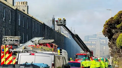 Firefighters using a height vehicle to battle a blaze, other members of the emergency services are in the street.