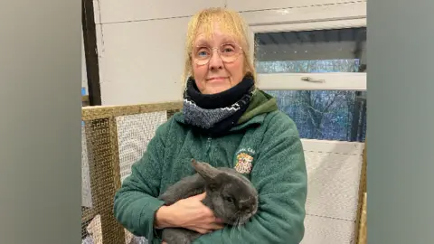 Animal Care A woman stands smiling at the camera holding a grey rabbit 