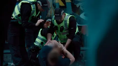 Several police officers in high-visibility vests restrain a person on the ground outside a building entrance. One officer kneels beside the individual while others stand nearby, with the scene partially framed by blurred objects in the foreground. The image is darkly lit, and police uniforms and equipment are clearly visible.