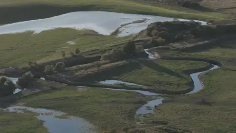 A river in a green fields, which swirls off course into more green fields in a bendy shape. 