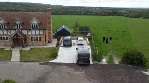 Jamie Niblock/BBC A drone photo of a farmhouse with a green farmers field behind it. There is also a driveway to the right of the farmhouse with three vehicles parked on it. There are three figures stood in the field
