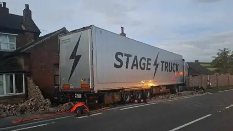 Supplied A grey lorry with the word "Stage Truck" on the side, which has left the road and brick rubble can be seen around it.