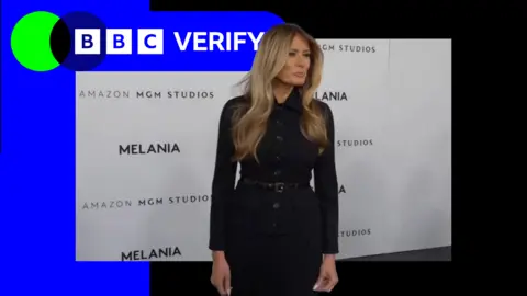 The First Lady of the United States Melania Trump stands at the premiere for her documentary Melania. She is wearing a long sleeved black dress and is standing in front of a banner that has 'Melania' written in black capital letters. 