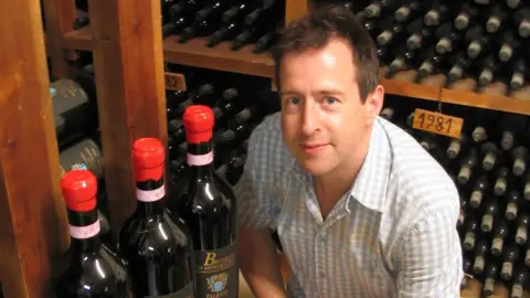 The Bell at Sapperton A photo of a white man with brown hair and wearing a chequered white and grey shirt. There is a rack of wine bottles behind him and three bottles in front of him