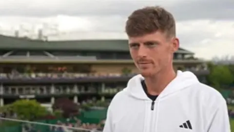 BBC Billy Harris wears a white zip-up jumper, he stands on a balcony at Wimbledon. 
