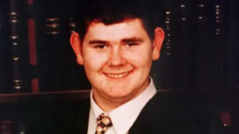 Family Handout Young man wearing a suit and tie standing in front of a bookcase smiling at the camera