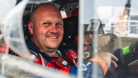James Williams Rallying Bald man smiling sitting in driving seat of car wearing race suit.