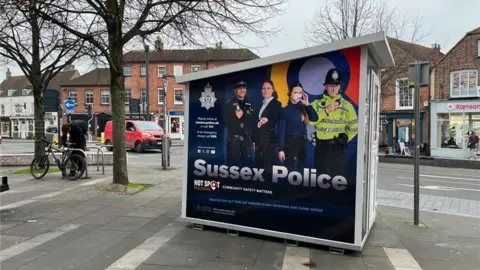 A police box in Chichester City Centre on a pavement with an advert for Sussex Police showing various uniformed and non uniformed officers