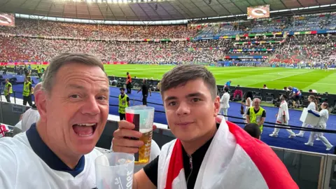 Arran Atkinson Arran left hand side taking a selfie with a cup of alchohol behind a football staduim backdrop. 