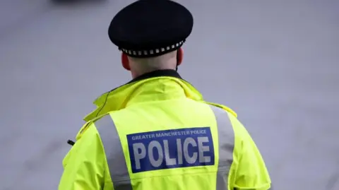 PA Media A police officer is standing with his back towards the camera. He is wearing a high-vis police jacket with the words "Greater Manchester Police" on the back and a black police hate.