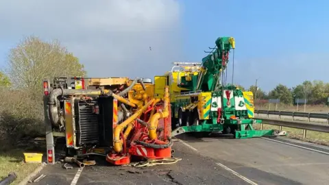 A brightly coloured lorry on its side on the left of a dual carriageway on the A11. It has a mix of orange and yellow pipes on its rear end. Just behind it on the other carriageway is a green and yellow recovery lorry