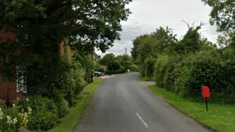 Murcot Turn, a road in Evesham, seen from the junction. There are grass verges and trees and houses along the road with flower beds. There is a post box, a parked car and a road sign.
