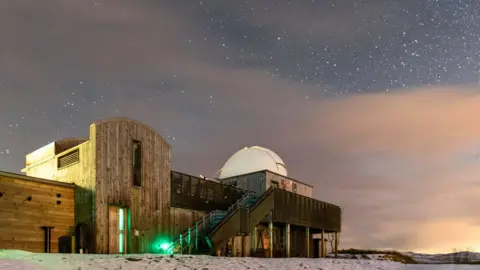 A wooden-fronted observatory building on snowy ground underneath a very starry sky