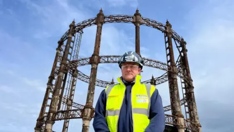 Andrew Turner/BBC Gavin Craig is standing in front of the Great Yarmouth gasometer, wearing a white hard hat, safety glasses, and a high visibility yellow tabbard over his blue jacket. Behind him is the gas holder, which is made from cast iron columns and lattice work, with finials on top of the columns. The sky behind is a grey/blue.