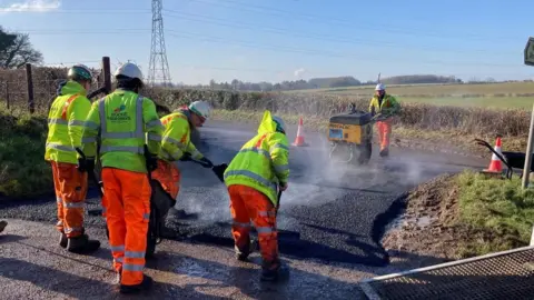 Wiltshire Council A group of workmen in fluorescent clothing on a roadside work to lay down tarmac. 