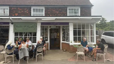 Lili Sheppard/BBC The exterior of Benenden's Community Shop in Benenden, Kent. A number of customers are sat outside eating and drinking. A sign saying "Benenden's 10th Anniversary" can be seen in the image.