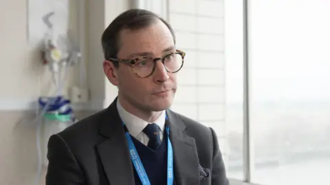 George Murphy stands in front of a large window and there is medical equipment on the wall behind him. He is wearing tortoiseshell rimmed glasses and a grey suit with a blue jumper underneath and a white shirt and dark blue tie.