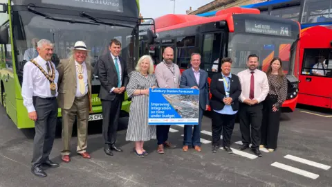 Nine people dressed smartly stand in a line in front of a series of buses at Salisbury Station forecourt.