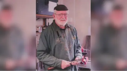 A man holding a tool and standing inside. He looks like he is woodworking.