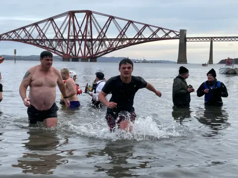 A man runs out of the Firth of Forth - he is dripping wet, wearing shorts and a T-shirt and has one eye closed. He appears to be running at pace.