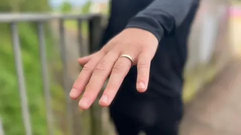 Network Rail A close-up of a man's hand with a gold wedding ring on it. The background is blurred and only the man's hand is in focus. In the background, the man's torso and legs are visible - he is wearing a black long-sleeved shirt and jeans, while standing on a railway bridge.