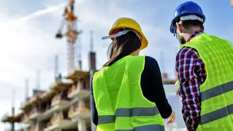 We see the backs of two people wearing high-vis green jackets over their clothes, standing in front of the blurred outline of a home being built with a crane overhead. The woman wears a yellow hard hat and has long dark hair. The man has a checked shirt on and a blue hard hat and a dark beard.