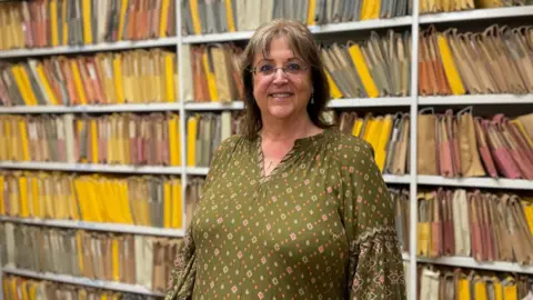 A woman with dark shoulder length hair and glasses smiles for the camera. She is wearing a dark green flowered top in front of a white shelving unit with colourful paper files