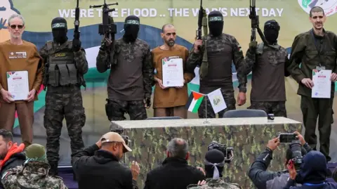 Masked gunmen holding weapons stand on stage with three men with white certificates