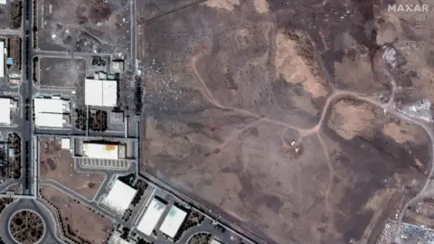 Reuters A black area with rubble strewn is  next to buildings in the dusty area of Natanz. A network of straight roads runs between the buildings, with a roundabout to the bottom left.