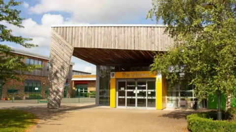 An entrance to a school building. Columns are painted yellow around a glass door, with a large wood pillar and roof, in front and above it respectively. Other buildings can be seen on the left and there is a tree on both the left and right of the photo. The words 'The Willows' are written above the door.
