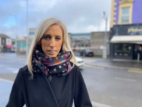 A woman with blonde hair down to her shoulders is standing in front of a street. She is wearing a scarf with blue, pink and orange stars. She is wearing a dark coat.