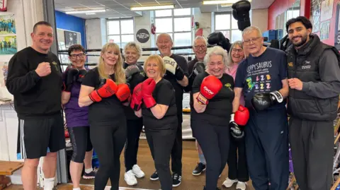 A group of elderly men and women dressed in dark gym gear hold up their boxing gloves, smiling.