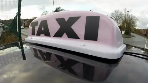 BBC A close-up of a taxi sign on top of a vehicle in a street.