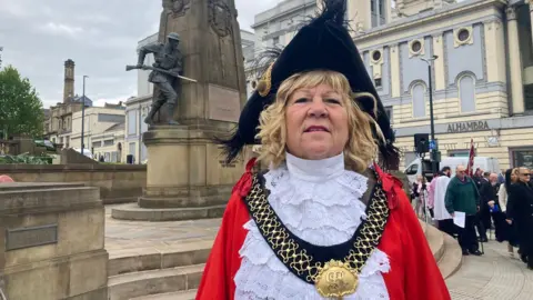 A woman dressed in red and white ceremonial robes with a gold mayoral chain and black tricorn standing in front of the Bradford War Memorial