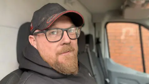 Man sat in a car with a black cap on and hoodie. He has a ginger beard and wears glasses.