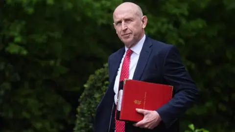 PA The minister is holding a red folder under his left arm as walks outside. He is dressed in a blue suit, white shirt and a red tie featuring white spots on it
