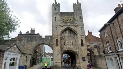 A medieval stone gate on York's city walls. A van drives under the left arch and people mill about in the background