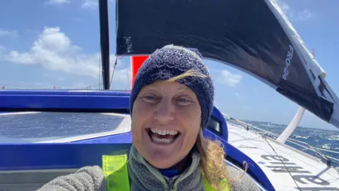 Pip Hare is wearing a blue hat and has long blonde hair. She is smiling. Behind her is a black sail and white hull - the ocean is behind the boat.