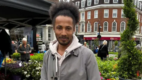 Josh is seen standing in front of a flower stall. He is wearing a grey jacket over a light-coloured hoodie.