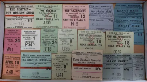 17 pastel-coloured ticket stubs laid out in a cardboard box for bands including The Beatles, The Searchers and Cliff Richard, all priced in shillings and pence.
