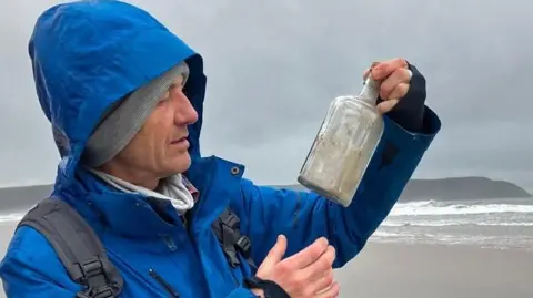 Mike Nicholls A man in a blue coat holding a bottle and looking at it. Behind him is a beach and sea. There is land in the background. The sky is grey and cloudy. 